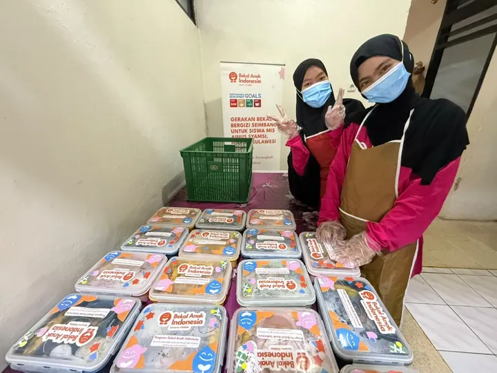 Packed lunches lined up on a table for the Bekal Anak Indonesia program, with two masked volunteers behind them
